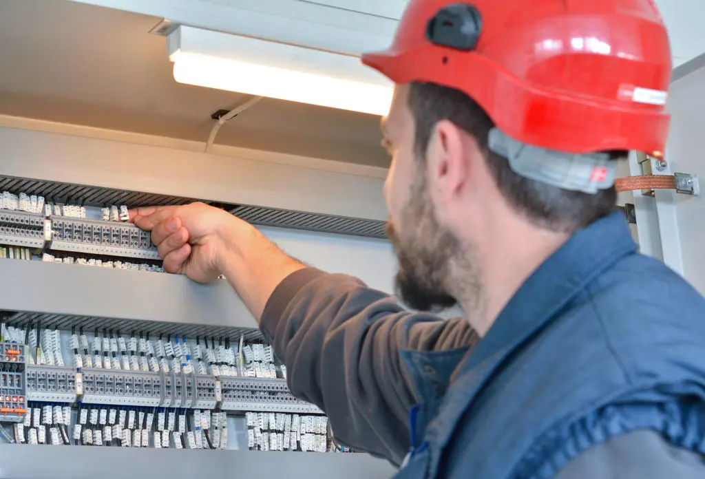 A man in red hard hat pointing to the bottom of an electrical panel.